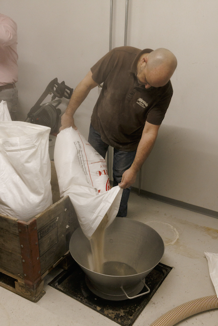 Ein Mann in Arbeitskleidung schüttet helle Whisky-Maische aus einem großen Sack in einen silbernen Behälter. Im Hintergrund sind Werkzeuge und weitere Säcke für den Macardo Brenn-Kurs zu sehen. Der Boden ist hell gefliest.